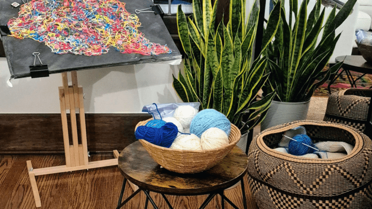 Colorful yarn art displayed on an easel beside a basket of yarns and indoor plants, illustrating a cozy crafting space.