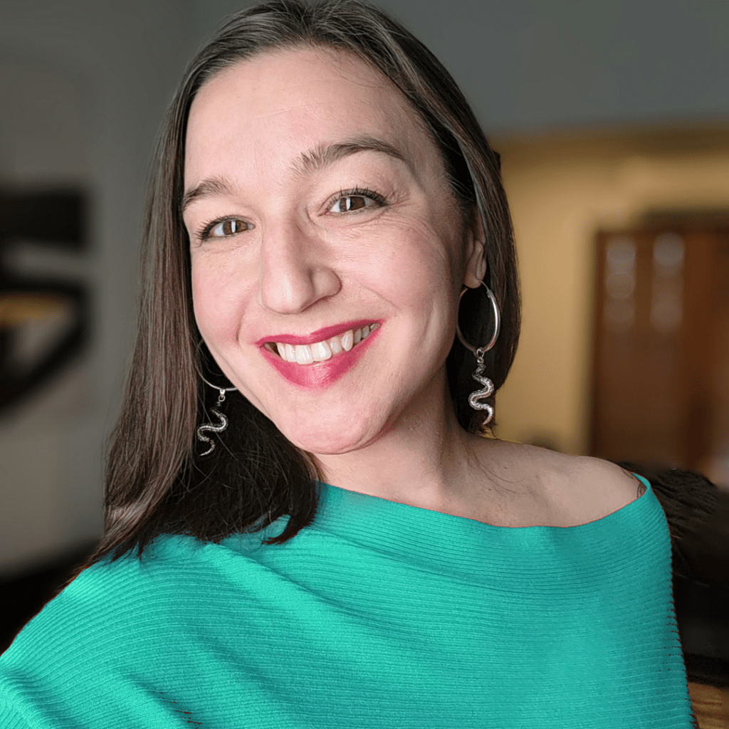 Woman with long dark hair smiles in a bright teal off-the-shoulder top and snake-shaped earrings. A soft background adds warmth to the image.