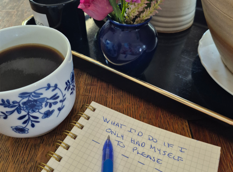 A cozy workspace featuring a cup of coffee, a blue vase with flowers, and an open notebook with handwritten thoughts on self-care.