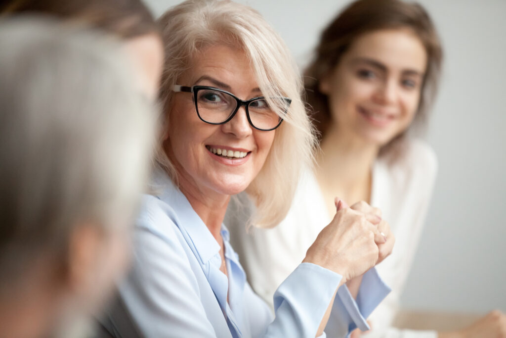 A smiling older woman with blonde hair and glasses engages in a meeting, surrounded by other participants, conveying a collaborative atmosphere.