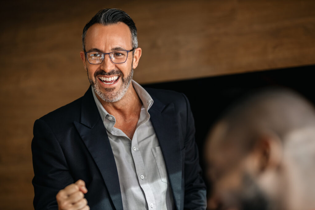 Smiling man in a suit expresses enthusiasm while gesturing, suggesting a dynamic discussion or presentation in a professional setting.