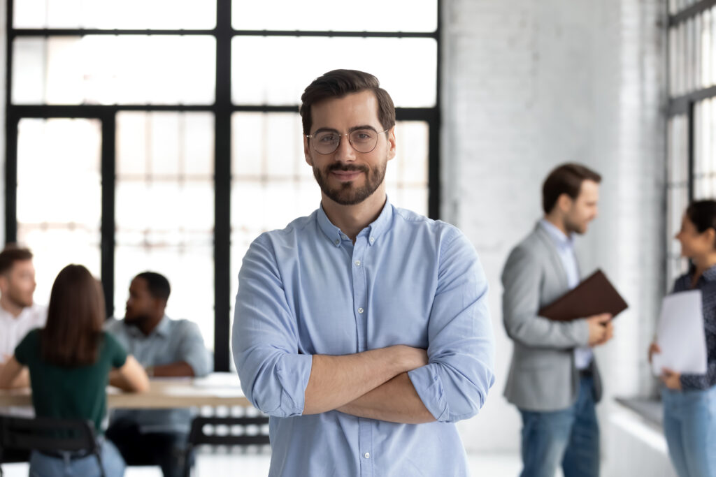Confident businessman in a light blue shirt stands with arms crossed, while diverse colleagues engage in discussion at a modern office setting.