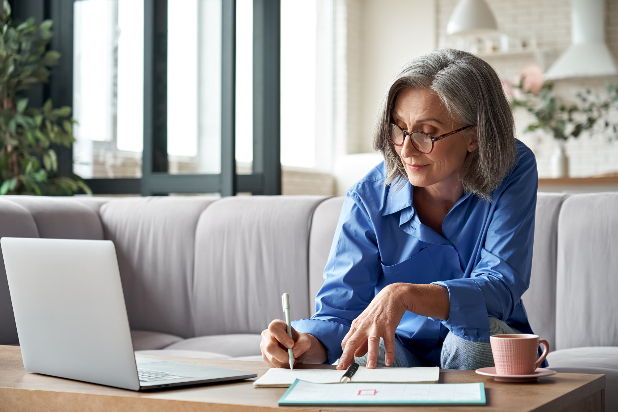 Older woman in a blue shirt writing in a notebook while working on a laptop at home. A coffee cup sits nearby, highlighting a productive workspace.