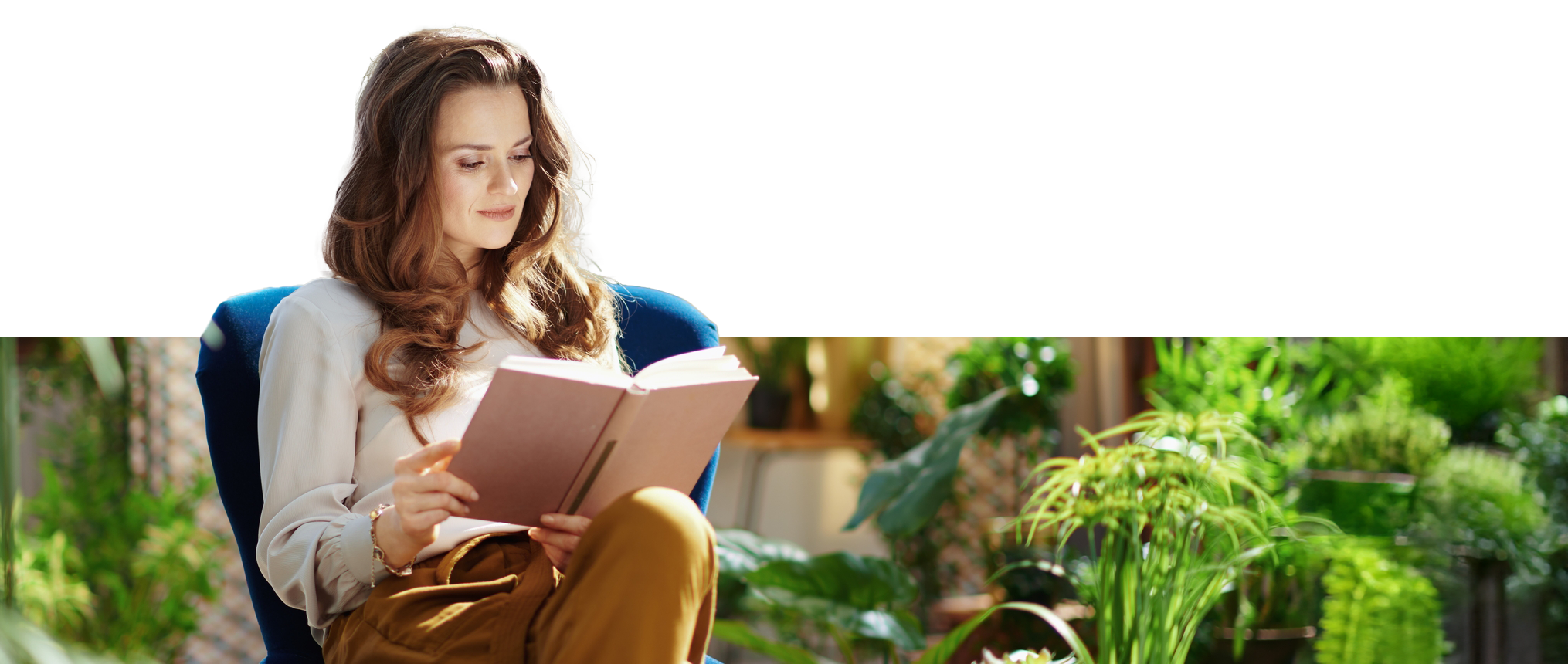 A woman with long wavy hair reads a book while seated in a blue chair, surrounded by lush indoor plants, conveying a peaceful, cozy atmosphere.