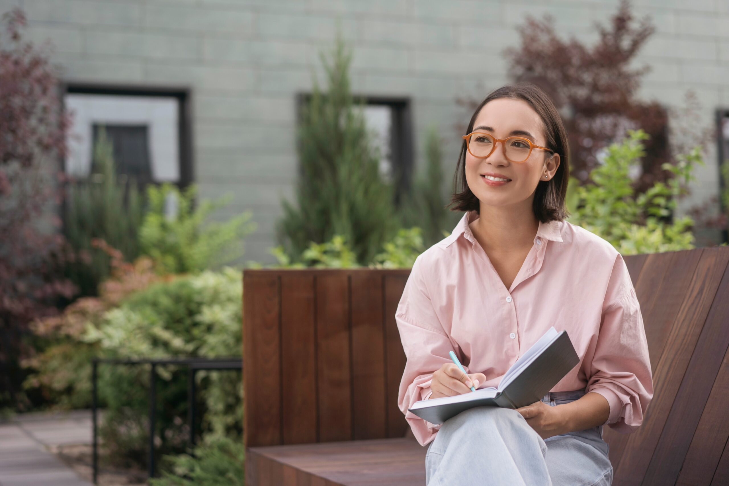 Portrait of confident smiling asian writer taking notes looking away sitting in park. Smart student wearing eyeglasses studying, reading book sitting in university campus. Education concept